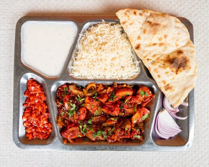A metal tray with sections of rice, naan, chicken chili, yogurt, chopped tomatoes, and red onion.