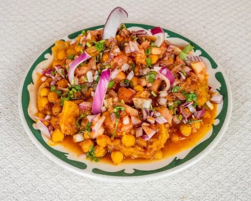 A vibrant plate of Indian chaat, topped with chickpeas, diced tomatoes, red onions, and cilantro.