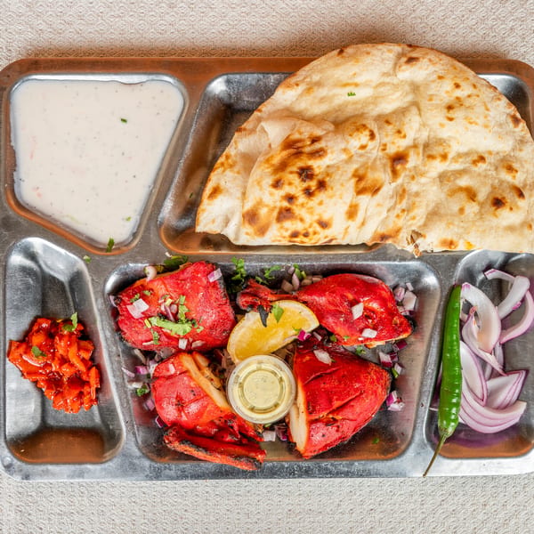 Indian thali with tandoori chicken, naan, raita, and pickled vegetables on a metal tray.