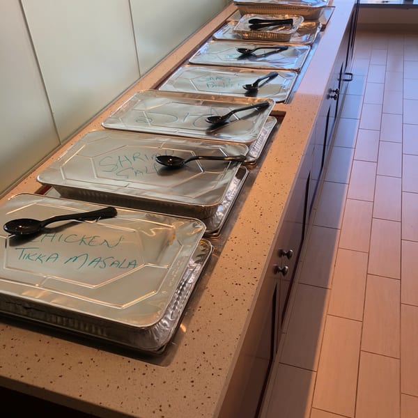 A buffet line of foil-covered catering trays with handwritten labels like "Chicken Tikka Masala" and "Saag Paneer".