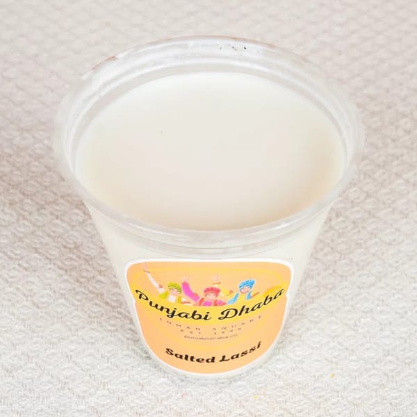 A clear plastic cup of white salted lassi from Punjabi Dhaba on a textured white surface.