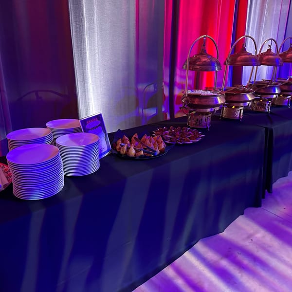 A buffet table with chafing dishes, plates, and appetizers under red and blue lighting.