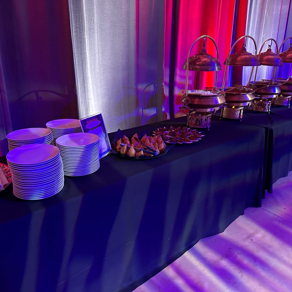 A buffet table with chafing dishes, plates, and appetizers under red and blue lighting.