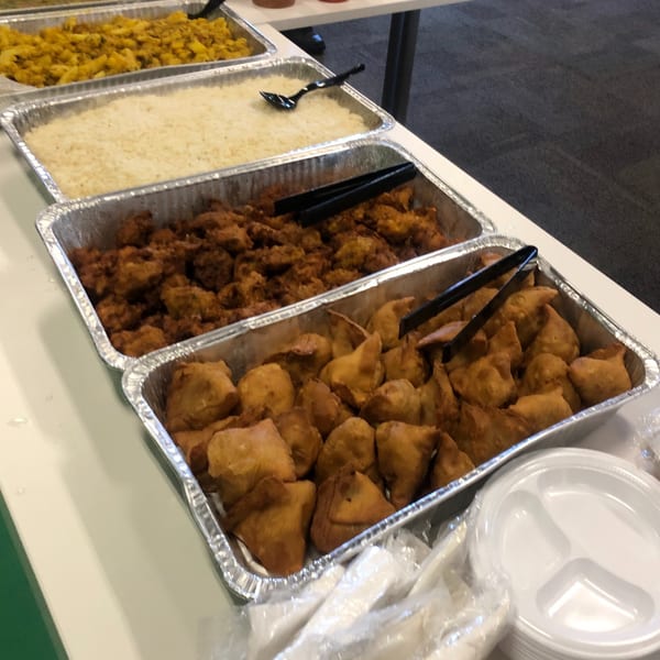 A buffet line with trays of Indian food, including samosas, rice, and curries, with drinks and plates.