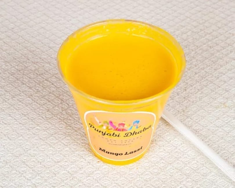 A yellow cup of mango lassi from Punjabi Dhaba with a white straw on a textured white surface.
