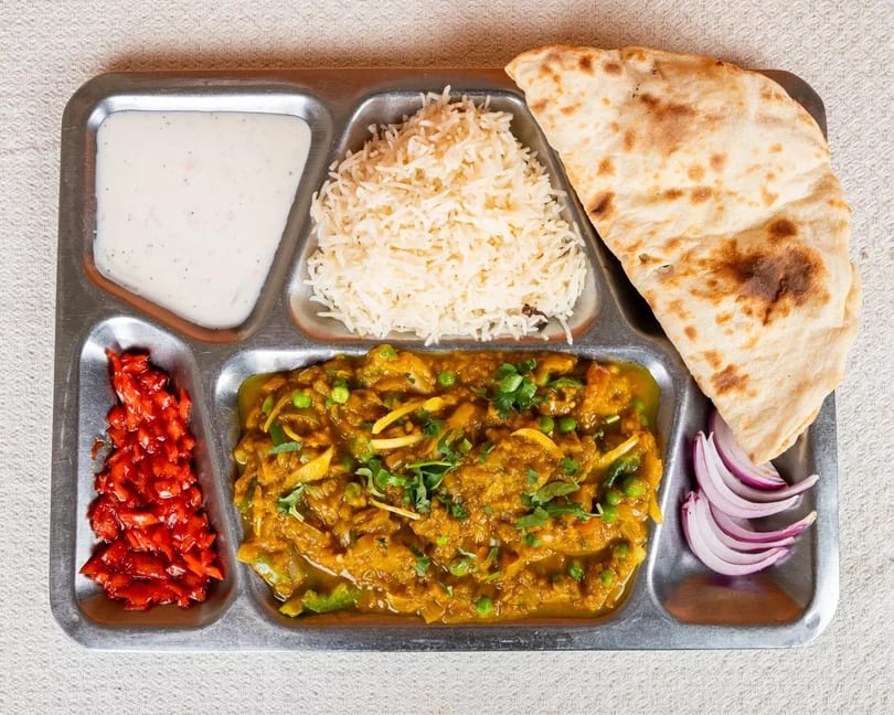 Indian thali with curry, rice, naan, raita, pickled vegetables, and red onion slices.