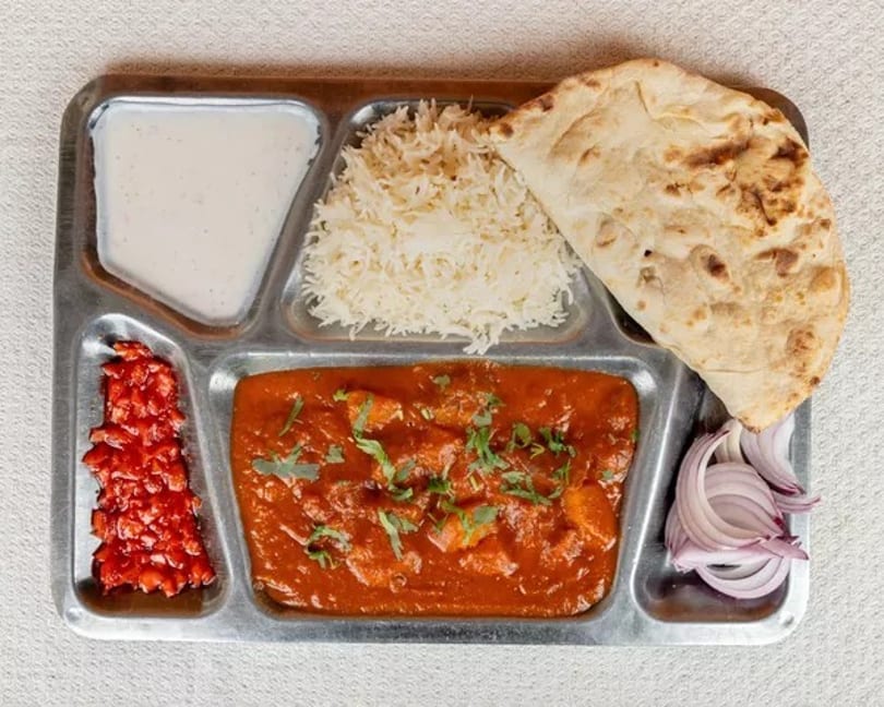 Indian thali with butter chicken, rice, naan, yogurt, pickled vegetables, and sliced onions.