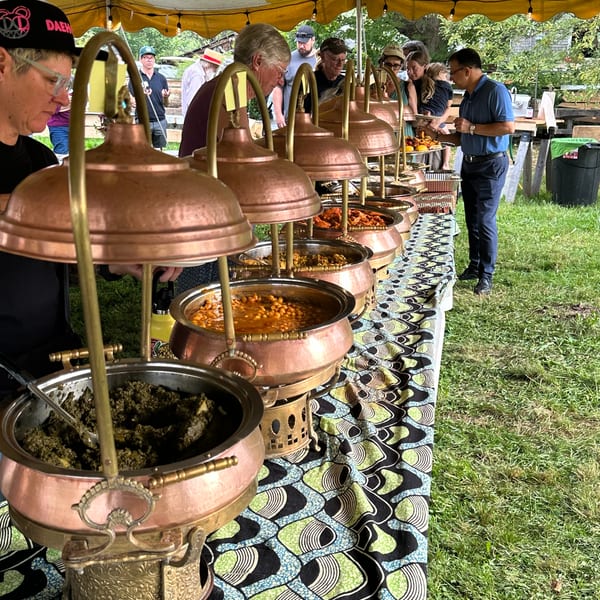 People serving themselves from a buffet line of copper chafing dishes under a striped tent.