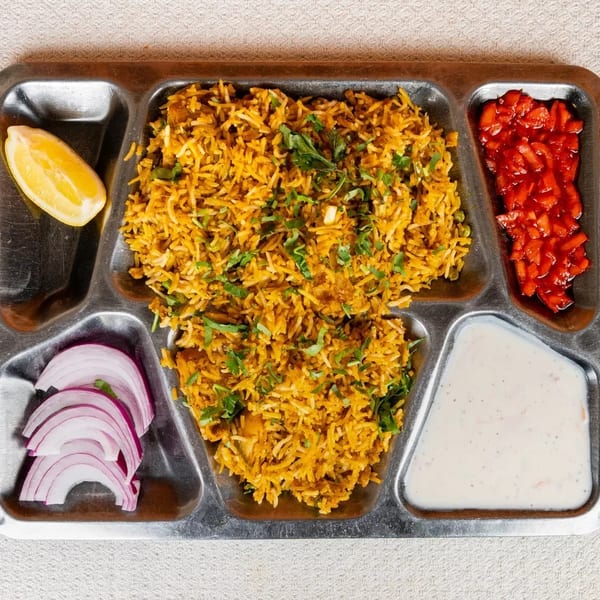 A metal tray with biryani, red onions, lemon, red relish, and a white sauce.