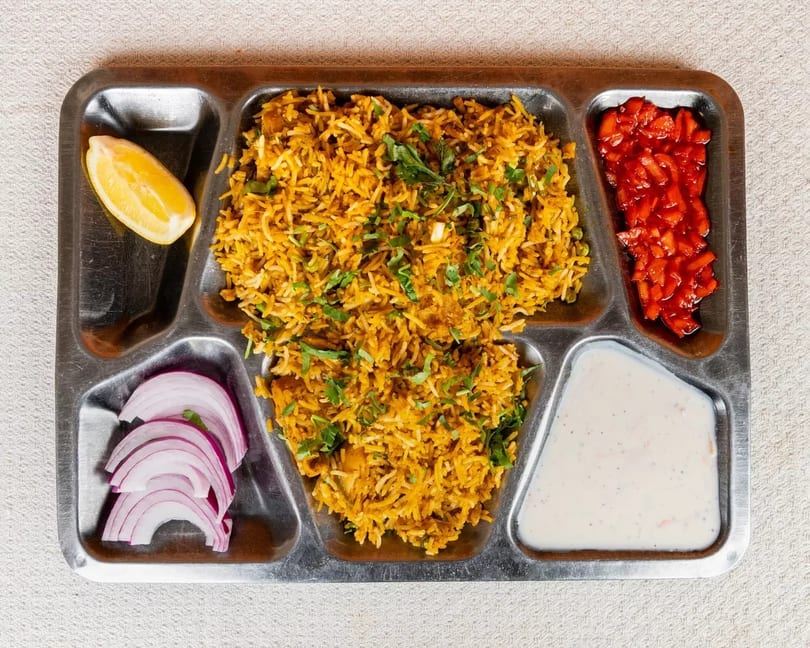 A metal tray with biryani, red onions, lemon, red relish, and a white sauce.